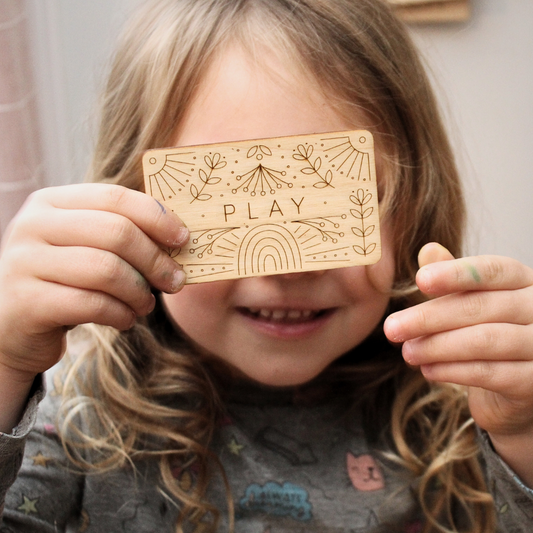 child holds a wooden word of the year card featuring the word 'play' along with intricately etched designs of flowers, a rainbow and other motifs. the word in the center is PLAY. 