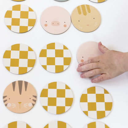 a grid of circular matching pieces are shown, some face-up, showing animals, some face-down showing yellow and white checks. a child’s hand reaches in from the right to pick up the pig pieces.