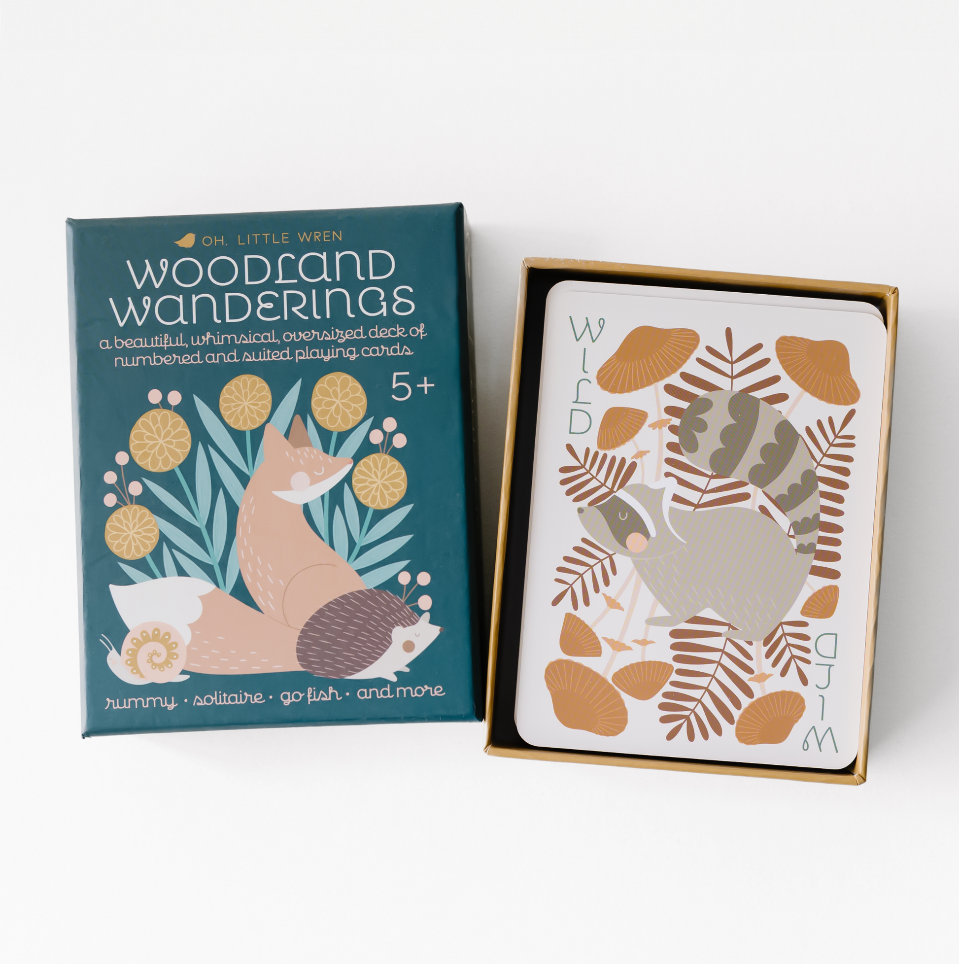 a box of woodland wanderings playing cards sits open, with the top on the lefthand side and the bottom on the righthand side--the bottom contains the stack of cards, with the wild card on top. the wild card has a raccoon surrounded by whimsical mushrooms.