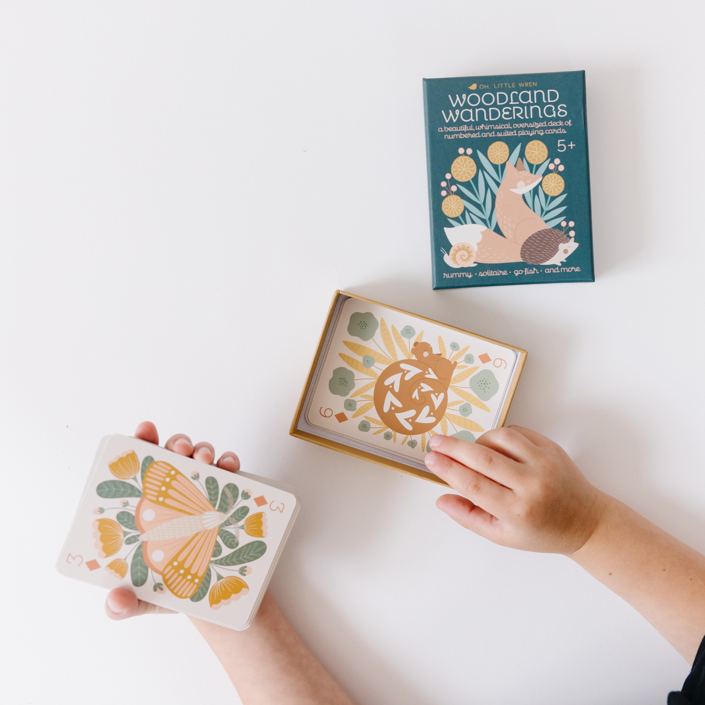 a child opens the box of woodland wandering playing cards. in one hand they hold the 3 of diamonds with a moth, and in the other the box bottom with a 6 of diamonds with a squirrel. 