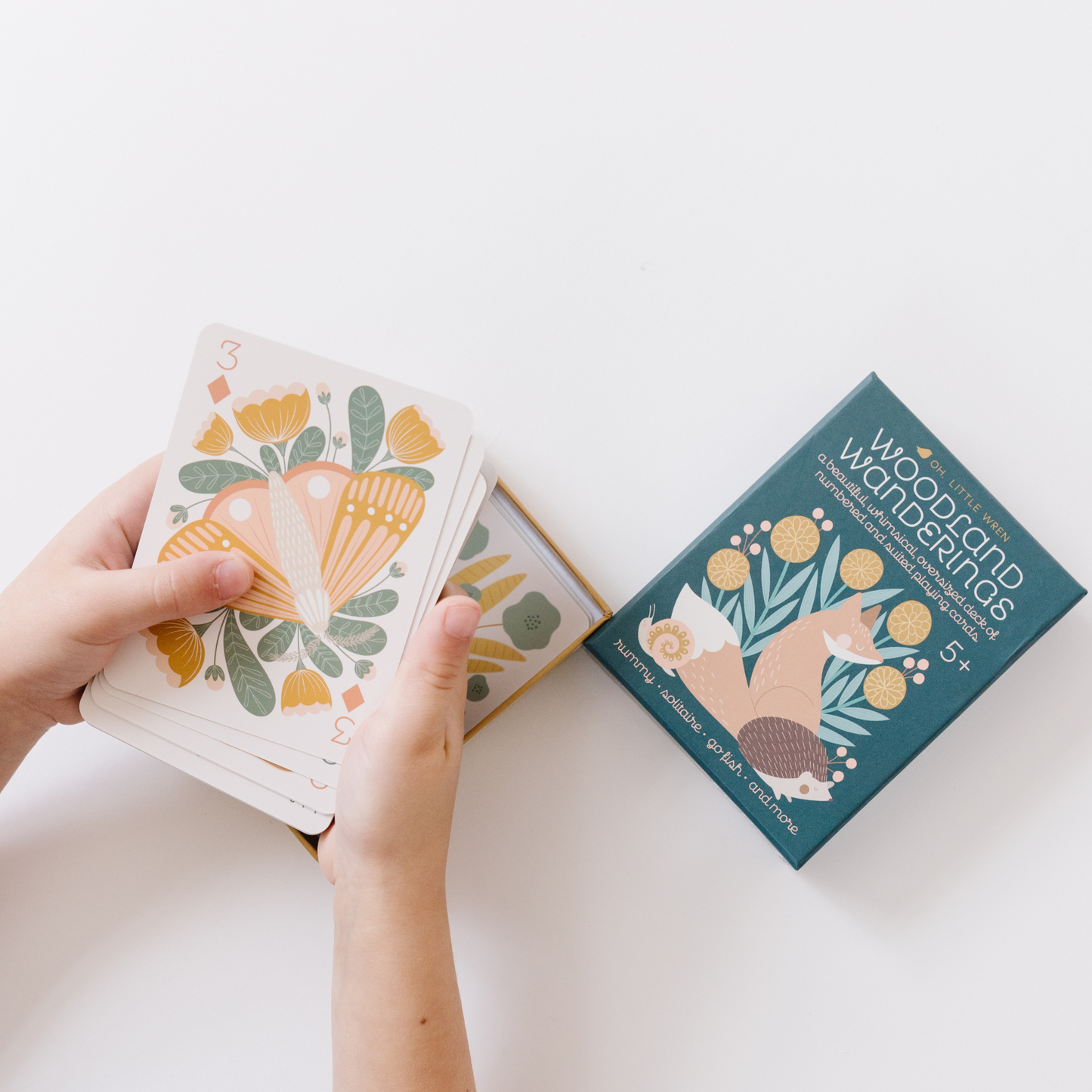 a child plays with the box of woodland wandering playing cards. they are holding the 3 of diamonds with a moth while the box lid sits to the right of their hands.