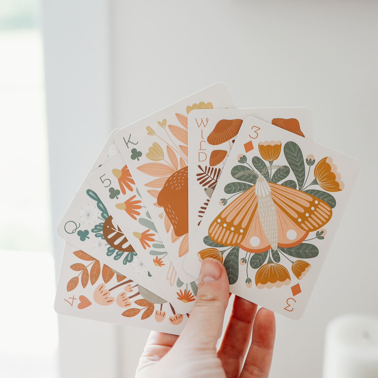 a hand holds 6 cards, fanned out. the front card, a 3 of diamonds, shows an intriccately illustrated moth surrounded by flowers.