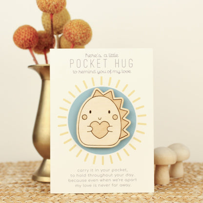 wooden dinosaur pocket hug is shown on its backer card propped up against a brass flower vase. card reads 'here's a little pocket hug to remind you of my love' and at the bottom 'carry it in your pocket, to hold throughout the day, because even when we're apart my love is never far away.'