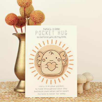 wooden axolotl pocket hugs is shown on its backer card propped up against a brass flower vase. card reads 'here's a little pocket hug to remind you of my love' and at the bottom 'carry it in your pocket, to hold throughout the day, because even when we're apart my love is never far away.'