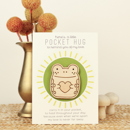 wooden frog pocket hug is shown on its backer card propped up against a brass flower vase. card reads 'here's a little pocket hug to remind you of my love' and at the bottom 'carry it in your pocket, to hold throughout the day, because even when we're apart my love is never far away.'