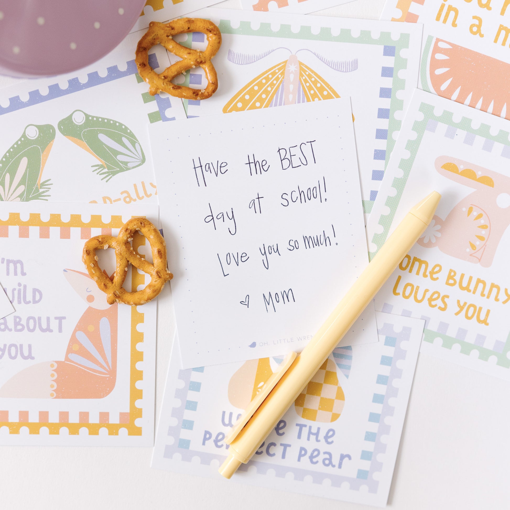 the back of the notecard is shown that includes the handwritten note 'have the BEST day at school! Love you so much! heart mom". more cards are in the background, as well as a yellow ballpoint pen and pretzels.