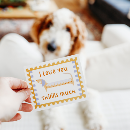 a lunchbox note with a picture of an exaggerately long dog reads 'i love you thiiiiiis much'. in the background there is an out of focus brown and white dog.
